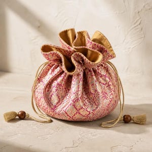 May include: A drawstring pouch featuring a vibrant magenta and gold floral brocade pattern. The bag has a gathered top, a solid gold-toned interior lining, and two gold cord drawstrings finished with dark brown wooden beads and gold tassels.