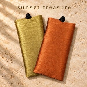 May include: Two rectangular fabric pouches, one in metallic gold and one in burnt orange, each featuring a textured surface and a small black knot closure at the top center. The text "sunset treasure" is printed above the pouches.