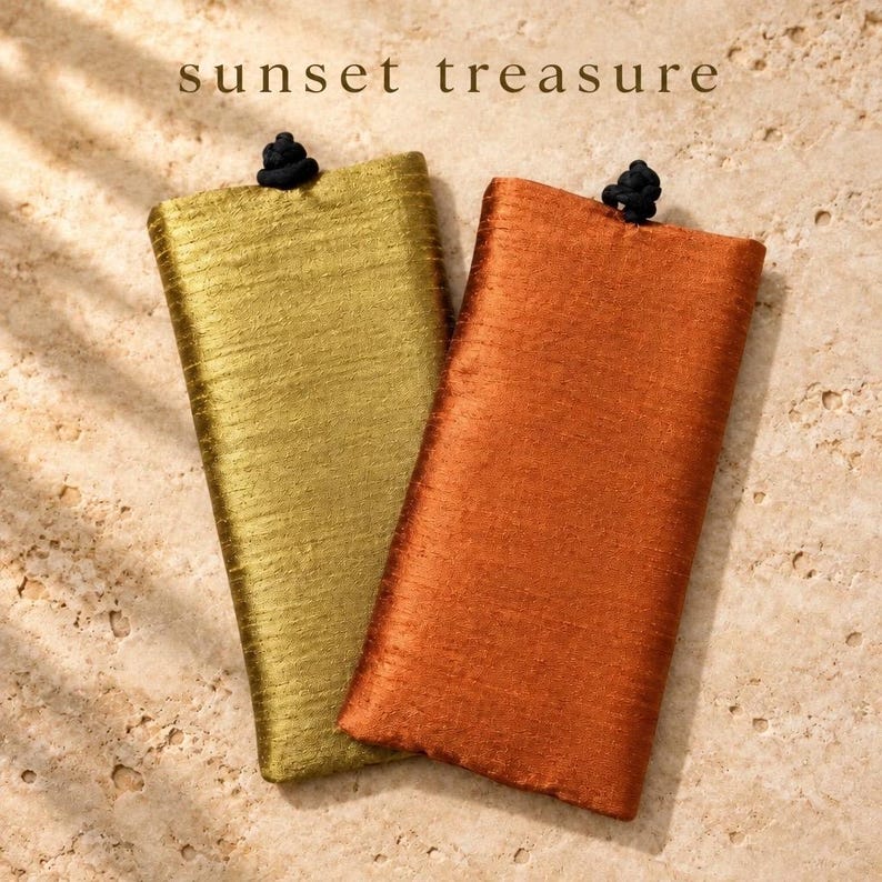 May include: Two rectangular fabric pouches, one in metallic gold and one in burnt orange, each featuring a textured surface and a small black knot closure at the top center. The text "sunset treasure" is printed above the pouches.