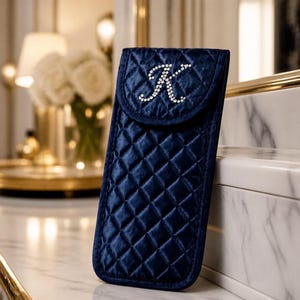 May include: A navy blue quilted fabric case with a diamond pattern and a flap closure. The front of the flap features a large, stylized letter K embellished with clear, sparkling rhinestones. The case has a vertical, rectangular shape.