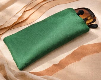 May include: A rectangular emerald green fabric pouch with a textured surface and a black drawstring closure. A pair of dark-rimmed sunglasses with brown lenses is partially tucked inside the pouch, with the frame visible at the top opening.