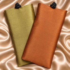 May include: Two rectangular fabric pouches, one in olive green and one in burnt orange, each featuring a smooth, slightly textured surface and a small black knotted cord closure at the top center edge of the rectangular opening.