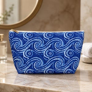 May include: A zippered pouch featuring a repeating pattern of blue and white swirling wave-like shapes. The design includes a cracked texture effect across the surface, with a gold-toned metal zipper pull visible at the top left corner of the bag.