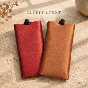 May include: Two rectangular fabric pouches, one in deep red and one in copper brown, each featuring a black knotted closure at the top center. The text "autumn ember" is printed in a simple font above the pouches.
