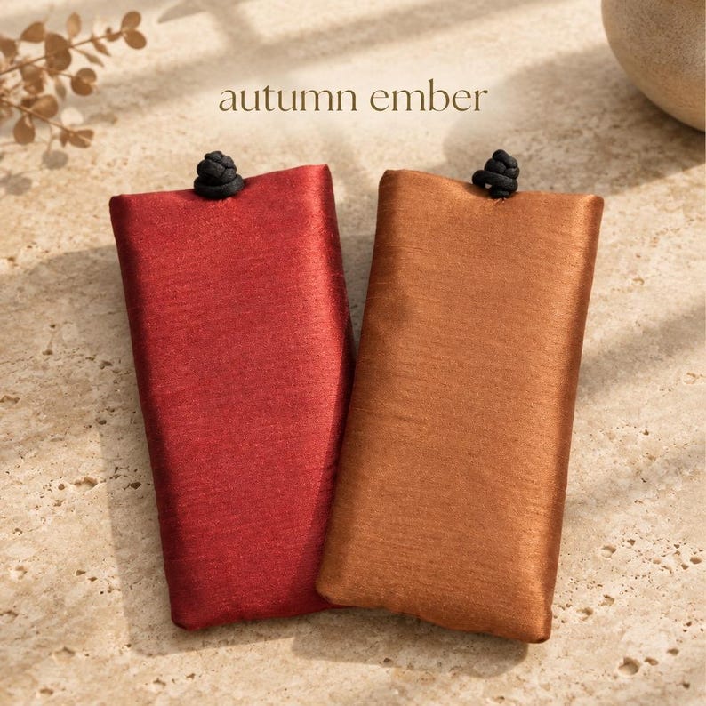 May include: Two rectangular fabric pouches, one in deep red and one in copper brown, each featuring a black knotted closure at the top center. The text "autumn ember" is printed in a simple font above the pouches.