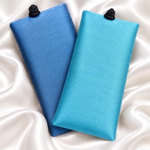 May include: Two rectangular fabric cases for glasses, one in deep blue and one in bright turquoise. Each case features a smooth, slightly shimmering texture and is topped with a small, round, black decorative knot closure at the center top edge.