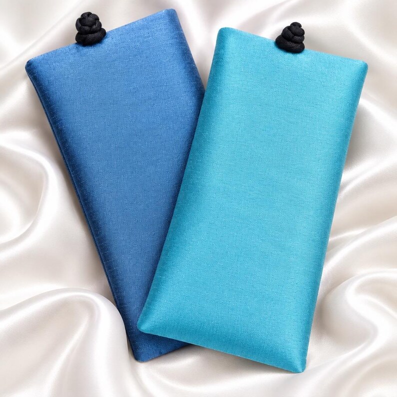 May include: Two rectangular fabric cases for glasses, one in deep blue and one in bright turquoise. Each case features a smooth, slightly shimmering texture and is topped with a small, round, black decorative knot closure at the center top edge.