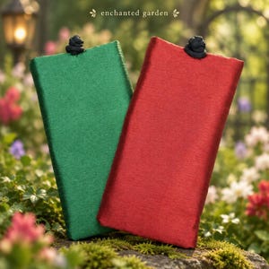 May include: Two rectangular fabric pouches, one in emerald green and one in deep red, each featuring a black knotted closure at the top center. The text "enchanted garden" is printed in small, elegant lettering above the pouches.