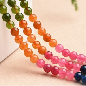 May include: A strand of colourful gemstone beads, featuring shades of pink, orange, green, and blue. The beads are round and smooth, and they are strung together on a clear thread.