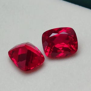 Może przedstawiać: Dwa fasetowane rubiny o szlifie poduszkowym. Kamienie szlachetne mają żywy czerwony kolor i są prezentowane na białej powierzchni w zielonym pudełku. Rubiny charakteryzują się wysokim stopniem przejrzystości i blasku.