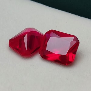 May include: Two faceted, bright red gemstones. One is a rectangular emerald cut, and the other is a smaller, geometric shape. The gems are displayed on a white surface inside a green box.