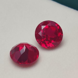 Könnte beinhalten: Zwei facettierte, ovale und runde, tiefrote Edelsteine. Die Edelsteine werden auf einer weißen Oberfläche in einer grünen Schachtel präsentiert. Die Edelsteine haben viele Facetten, die das Licht reflektieren.