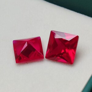 May include: Two square-cut, faceted, deep red gemstones. The gems are displayed on a white surface inside a green box. The gemstones have a brilliant, reflective quality, suggesting they are of high quality.