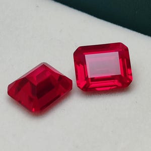 May include: Two faceted, rectangular, and diamond-shaped red gemstones. The gems are a vibrant, deep red color and appear to be cut with precision. They are displayed on a white surface within a dark green box.