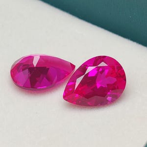 Puede incluir: Dos gemas rosas brillantes, facetadas en forma de pera, se exhiben sobre una superficie blanca dentro de una caja verde azulado. Las gemas tienen múltiples facetas que reflejan la luz, creando un efecto brillante. Las gemas son probablemente rubíes o piedras preciosas similares.