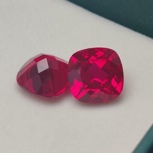 May include: Two faceted, vibrant red gemstones. One is cushion-cut, the other is a triangular shape. The gems are displayed in a white box with a dark green border. The gems have a high degree of clarity and brilliance.
