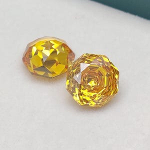 Peut inclure: Deux pierres précieuses jaunes à facettes, taillées en brillant. L'une est de forme ovale, l'autre est ronde. Les pierres sont présentées sur une surface blanche, avec un fond vert d'eau.