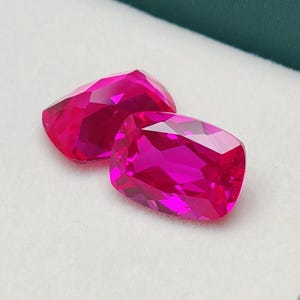 Może przedstawiać: Dwa fasetowane, szlifowane w kształcie poduszki, jaskraworóżowe kamienie szlachetne spoczywają w białym pudełku z zieloną obwódką. Kamienie mają wysoki połysk i odbijają światło, ukazując swoje skomplikowane szlify. Kamienie te są prawdopodobnie przeznaczone do tworzenia biżuterii lub kolekcjonowania.