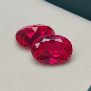 Puede incluir: Dos gemas rojas profundas, facetadas y de forma ovalada. Las gemas se exhiben en una caja blanca con un borde verde. Las gemas tienen un alto brillo y reflejan la luz, mostrando su talla y claridad. Las piedras preciosas son probablemente rubíes.