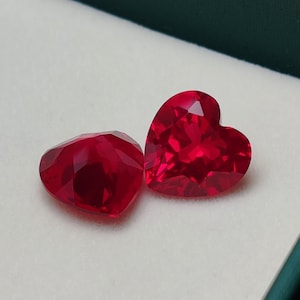 Può includere: Due pietre preziose rosse a forma di cuore, sfaccettate. Le gemme sono esposte su una superficie bianca all'interno di una scatola verde. Le sfaccettature catturano la luce, creando un effetto scintillante. Le gemme sono probabilmente rubini.