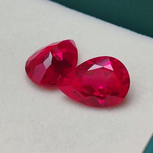 May include: Two faceted, pear-shaped, bright red gemstones. The gems are displayed on a white surface inside a green box. The gems have a high luster and reflect light, showcasing their cut and clarity.