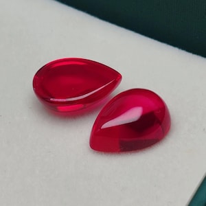 May include: Two polished, teardrop-shaped red gemstones are presented in a white box with a green border. The gems have a high-gloss finish, reflecting light. The gems are likely rubies or similar precious stones.