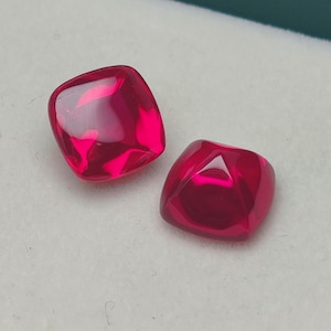 Op de afbeelding: Twee kussenvormig geslepen, gefacetteerde edelstenen in een levendige, dieprode tint. De edelstenen worden gepresenteerd op een wit oppervlak, met een vleugje groene achtergrond. De stenen hebben een gepolijst, reflecterend oppervlak.