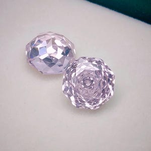 Może przedstawiać: Dwa fasetowane, jasnofioletowe kamienie szlachetne. Jeden ma kształt owalny, drugi okrągły, przypominający kwiat. Kamienie są umieszczone na jasnym tle z ciemnozieloną obwódką.