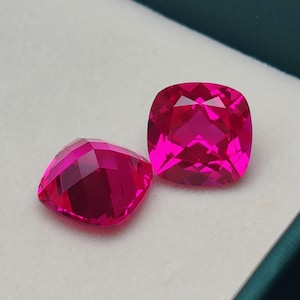 May include: Two faceted, cushion-cut gemstones in a vibrant magenta hue. The gems are displayed on a white surface within a dark green box, showcasing their brilliant cut and color. These gems are ideal for jewelry making.