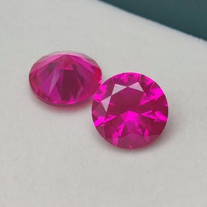 May include: Two round, faceted, hot pink gemstones. The gems are displayed on a white surface inside a teal box. The gems have many facets that reflect light, creating a sparkling effect. The gems are a vibrant, saturated pink color.