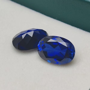 Puede incluir: Dos gemas ovales facetadas de color azul oscuro. Las gemas se muestran sobre una superficie blanca dentro de una caja verde. Las piedras tienen un corte brillante, reflejando la luz y creando un efecto brillante. Adecuado para joyería.