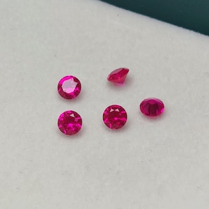 May include: A collection of five round, faceted, bright pink gemstones. The gems are displayed on a white surface, with a hint of a green border in the background. The gemstones are likely rubies, perfect for jewelry making.