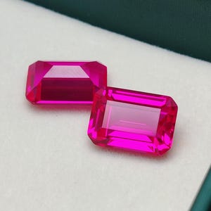 May include: Two vibrant, rectangular-cut, hot pink gemstones. The gems are set against a white background inside a dark green box. The facets of the stones reflect light, showcasing their clarity and vibrant colour.