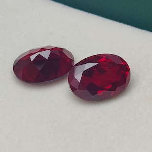 Op de afbeelding: Twee ovaal geslepen, gefacetteerde edelstenen met een dieprode kleur. De edelstenen worden gepresenteerd in een wit, met fluweel gevoerd doosje met een groene rand. De facetten vangen het licht op, waardoor een sprankelend effect ontstaat.