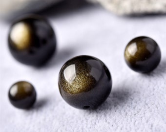 Könnte beinhalten: Vier runde, schwarze Perlen mit goldenen Sprenkeln. Die Perlen sind glatt und glänzend.