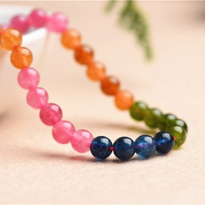 May include: A multi-coloured beaded bracelet with pink, orange, green, and blue beads. The bracelet is made of natural stones and is strung on a red thread.