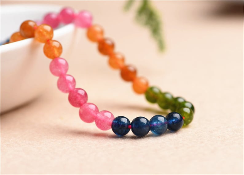 May include: A multi-coloured beaded bracelet with pink, orange, green, and blue beads. The bracelet is made of natural stones and is strung on a red thread.