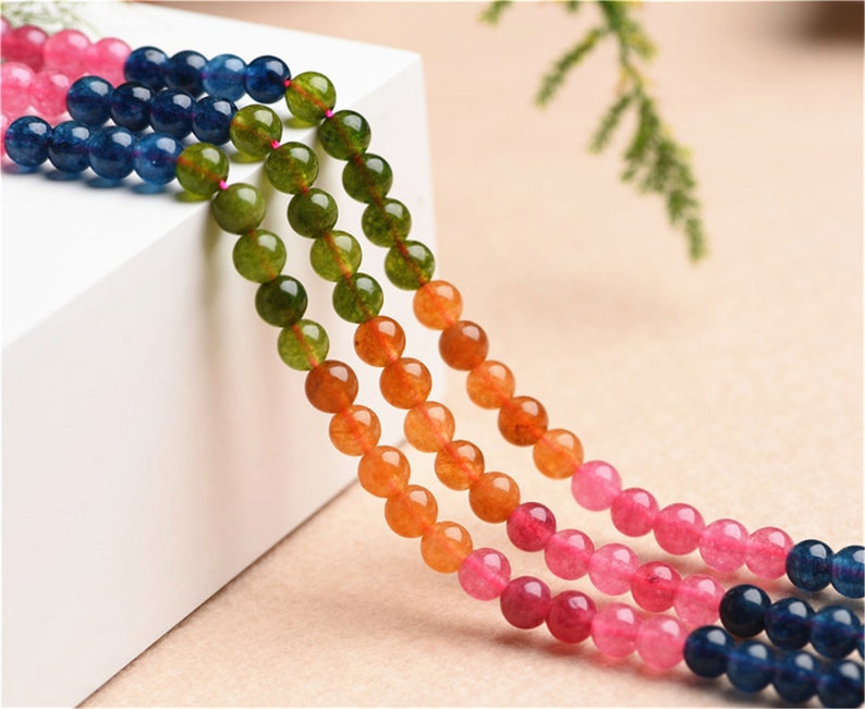 May include: A strand of colourful gemstone beads, featuring shades of pink, orange, green, and blue. The beads are round and smooth, and they are strung together on a clear thread.