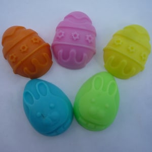 May include: Five Easter-themed egg-shaped soaps. Two are decorated with a pattern of stars and stripes, one is orange, one is yellow, and one is pink. The other two soaps are green and blue and feature a bunny rabbit design.