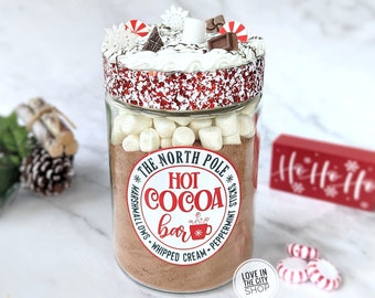 May include: A clear glass jar filled with hot cocoa mix, marshmallows, and toppings. The jar is labeled "The North Pole Hot Cocoa Bar" and is decorated with red and white accents, including peppermint candies and chocolate pieces. A festive holiday treat.