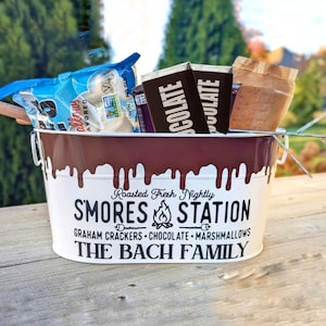 May include: A white metal bucket with a brown chocolate-like drip design. The bucket has a black and white graphic that says "Roasted Fresh Nightly SMORES STATION GRAHAM CRACKERS - CHOCOLATE - MARSHMALLOWS THE BACH FAMILY". The bucket is filled with graham crackers, chocolate bars, and marshmallows.