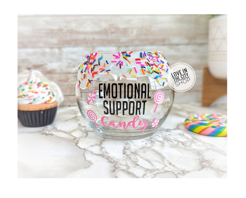 May include: A clear glass jar with a white rim and sprinkles around the top. The jar has a pink and black sticker that says "Emotional Support Candy".