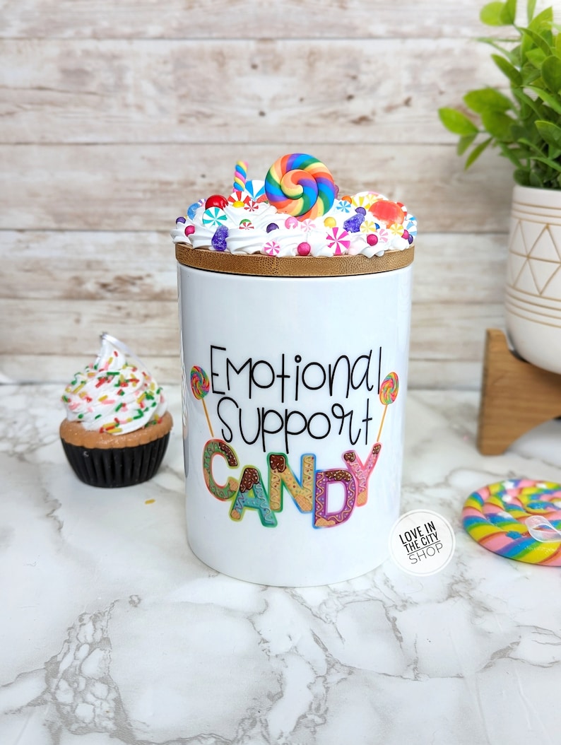 May include: White ceramic jar with a bamboo lid, decorated with a candy-themed design. The jar features the text "Emotional Support Candy" in colorful lettering. The lid is topped with a swirl of white frosting and various candies, including a rainbow lollipop.