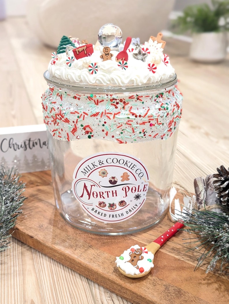 May include: A clear glass jar with a lid decorated with faux white frosting, miniature gingerbread figures, and holiday charms. The rim features silver glitter and red and green sprinkles. A label reads Milk & Cookie Co. North Pole Baked Fresh Daily.