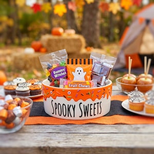 May include: A white ceramic bowl with an orange drip pattern, colorful sprinkles, and the text "SPOOKY SWEETS" printed in black. The bowl is filled with various packaged candies, including Gummy Witches, a ghost-themed bag, and fruit chews.
