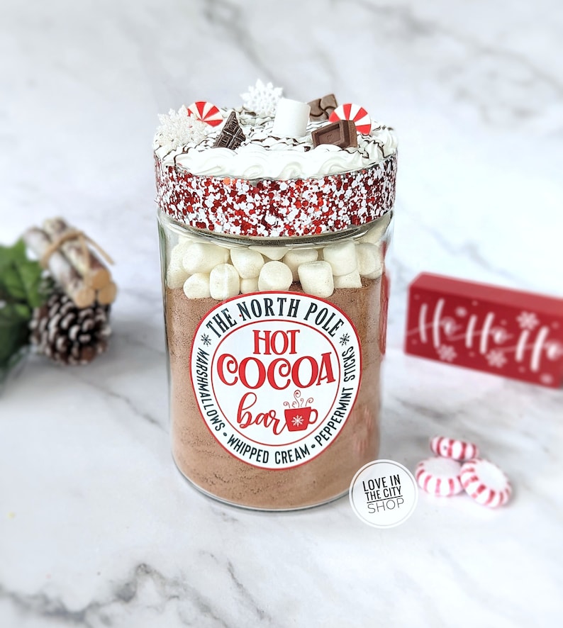 May include: A clear glass jar filled with hot cocoa mix, marshmallows, and toppings. The jar is labeled "The North Pole Hot Cocoa Bar" and is decorated with red and white accents, including peppermint candies and chocolate pieces. A festive holiday treat.