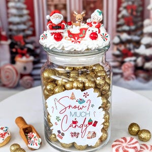 May include: A glass jar filled with gold-colored candies, topped with a decorative Christmas scene featuring Santa Claus, Mrs. Claus, and a reindeer. The jar has a label that reads "Snow Much Candy."
