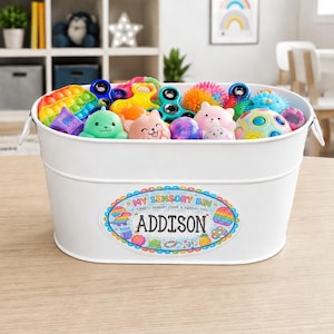 May include: A white metal bin filled with colorful sensory toys, including fidget spinners and squeeze toys. The bin has a label that reads "My Sensory Bin" and the name "Addison".
