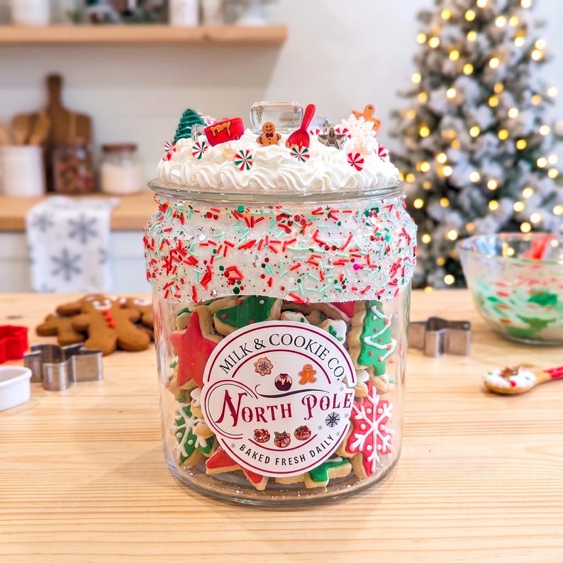 May include: A clear glass jar filled with assorted holiday-shaped cookies, featuring a label that reads Milk & Cookie Co. North Pole Baked Fresh Daily. The lid is decorated with white faux frosting, red and green sprinkles, and miniature ornaments.