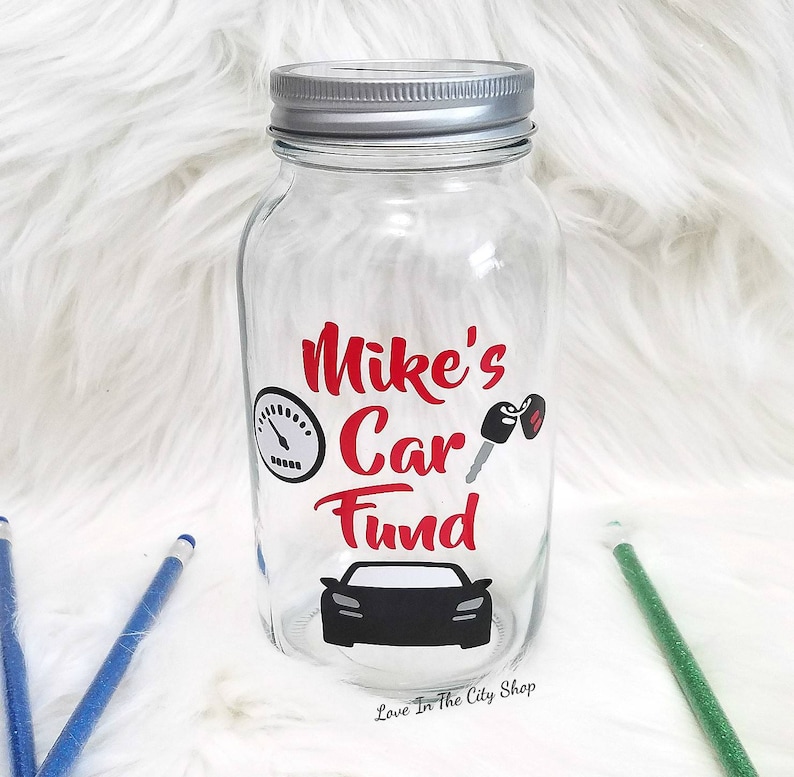 Op de afbeelding: Een glazen pot met een zilveren deksel. De pot heeft een rood en zwart ontwerp met de tekst "Mike's Car Fund" met een snelheidsmeter, autosleutels en een zwarte auto afbeelding. De pot staat op een wit, pluche oppervlak.